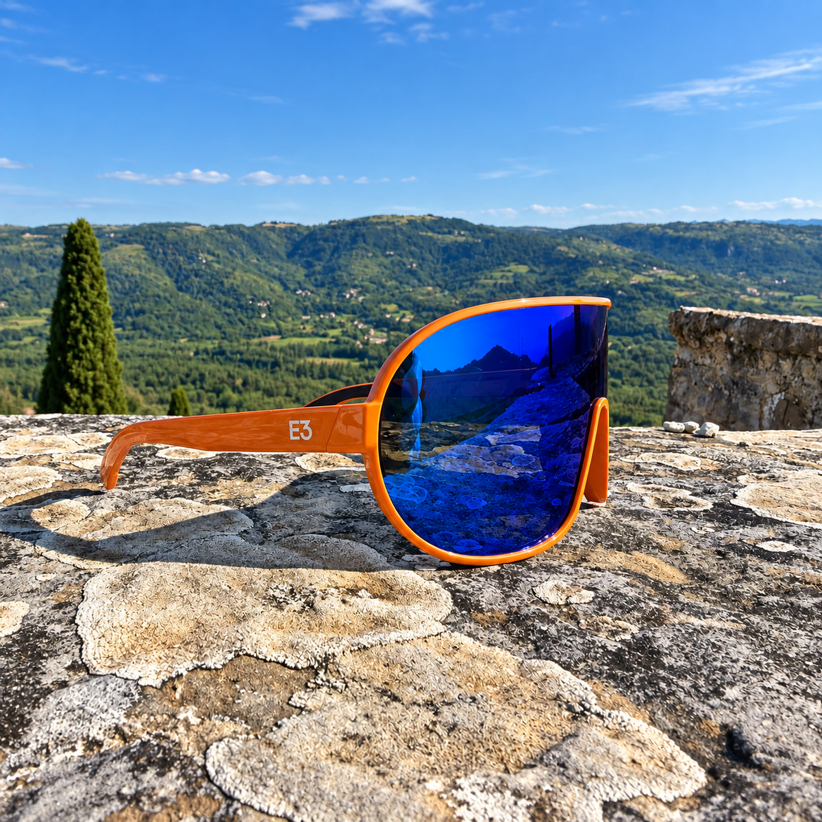 Large round sunglasses with orange wood-look frames and mirrored blue lenses, resting on a stone surface with a scenic mountain landscape and blue sky in the background.