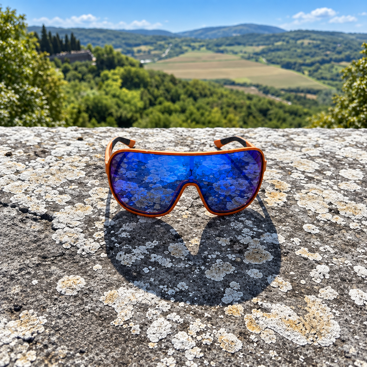 Orange wrap-around sunglasses with reflective blue mirrored lenses resting on a textured stone wall, with a scenic green valley and hills under a blue sky in the background.