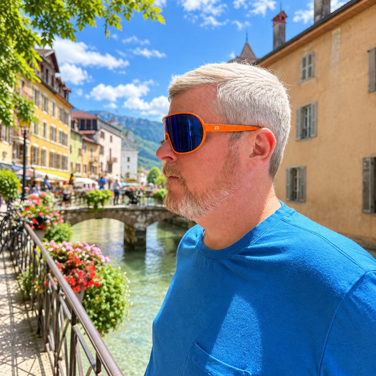 A man with white hair and a beard wearing bright orange sunglasses and a blue t-shirt, standing outdoors next to a canal in a European town with colorful buildings and flowers.