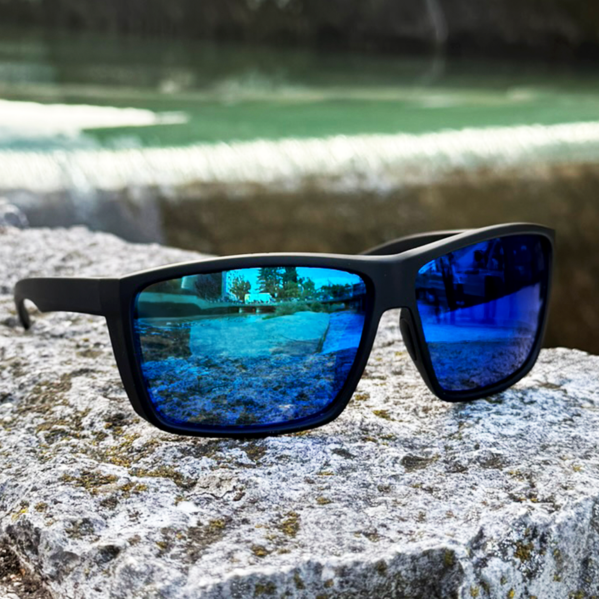 E3 Optics E1-B1 blue mirror polarized sunglasses with soft-touch black frame resting on a stone ledge beside turquoise water — ideal for boating and fly fishing — angled close-up view.
