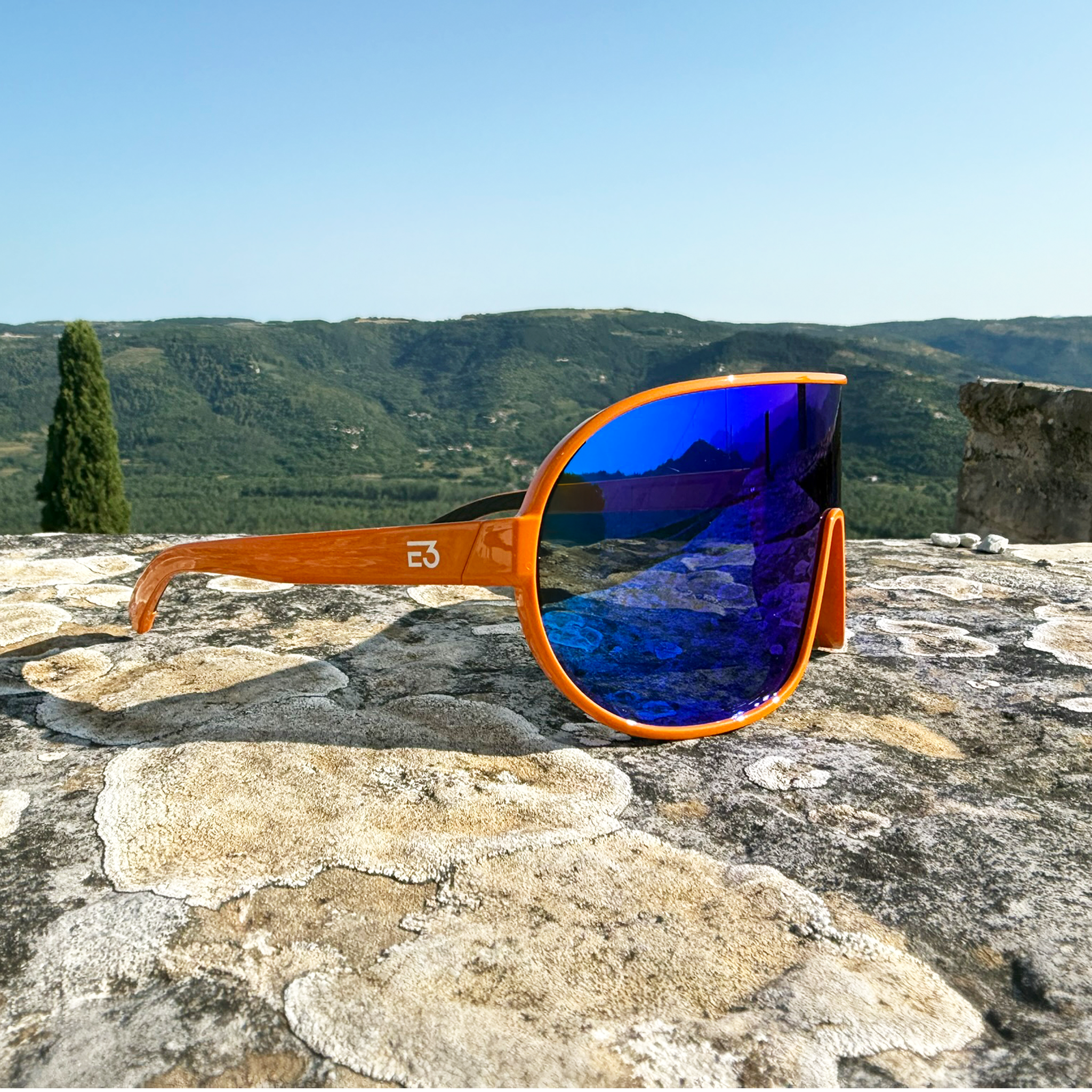 E3 Optics E5 sunglasses in orange frame with blue mirror lens resting on a stone wall overlooking rolling green hills.