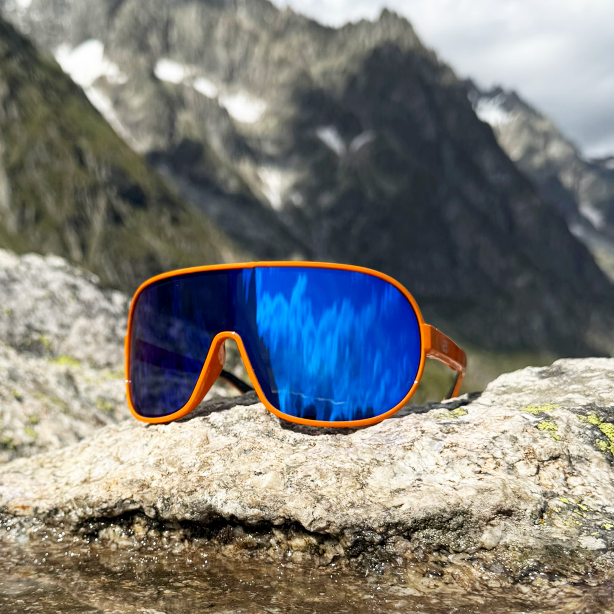 E3 Optics E5 sunglasses featuring orange frame and blue mirror lens placed on a railing with cloudy mountain scenery.