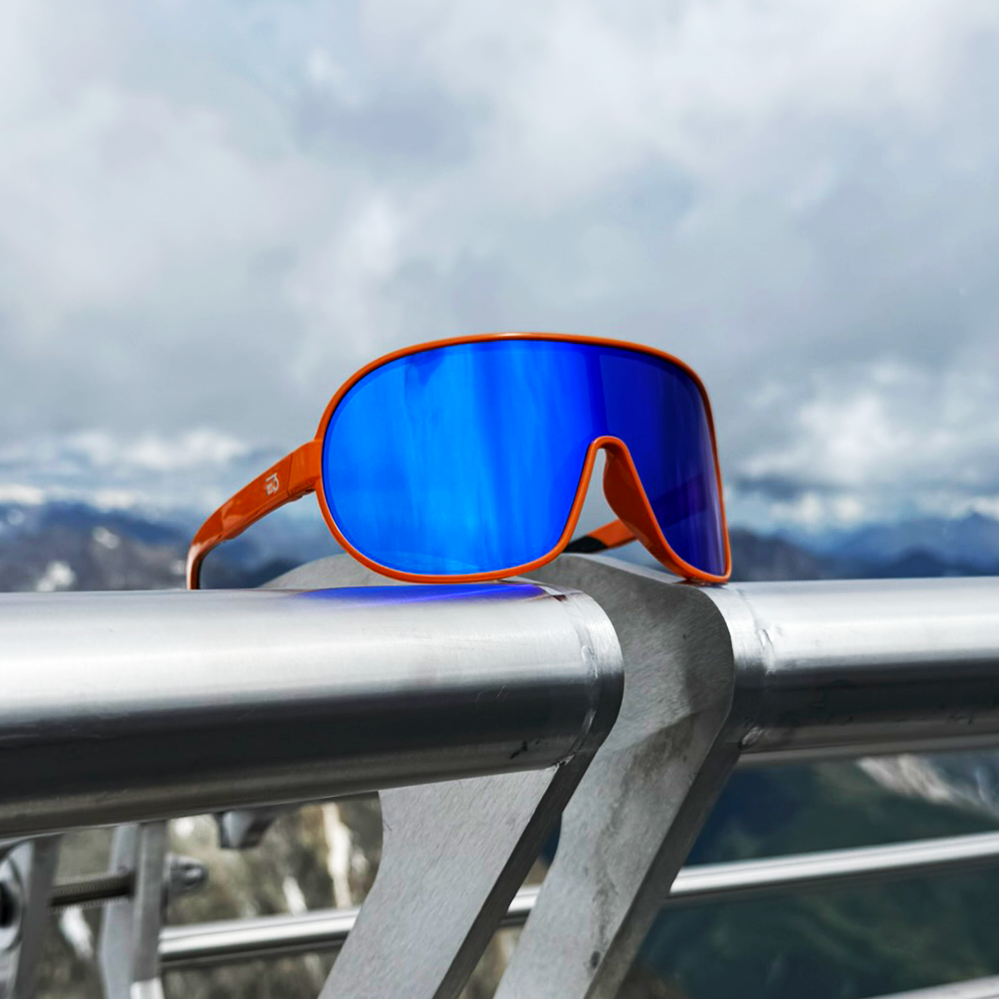 E3 Optics E5 sunglasses with orange frame and blue mirror lens resting on a rock with alpine mountains in the background.