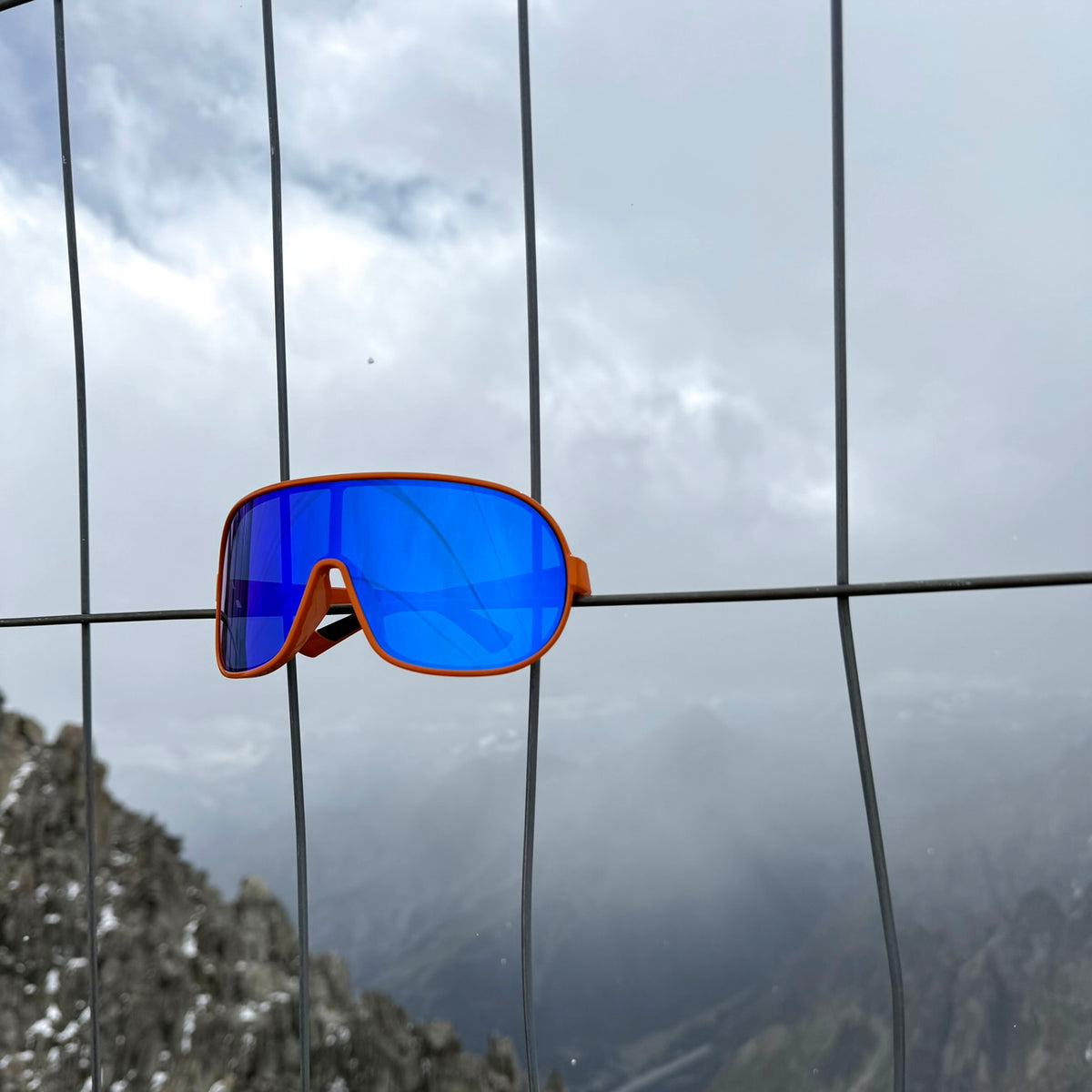 Orange-framed sport sunglass with a vibrant blue mirrored lens hanging on a wire fence, with a cloudy mountain landscape in the background.s