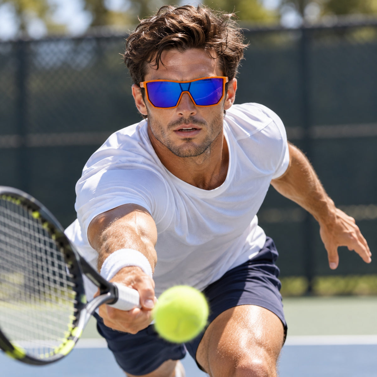A man wearing orange-framed mirrored sunglasses and a white t-shirt plays tennis on an outdoor court, reaching for a yellow tennis ball.