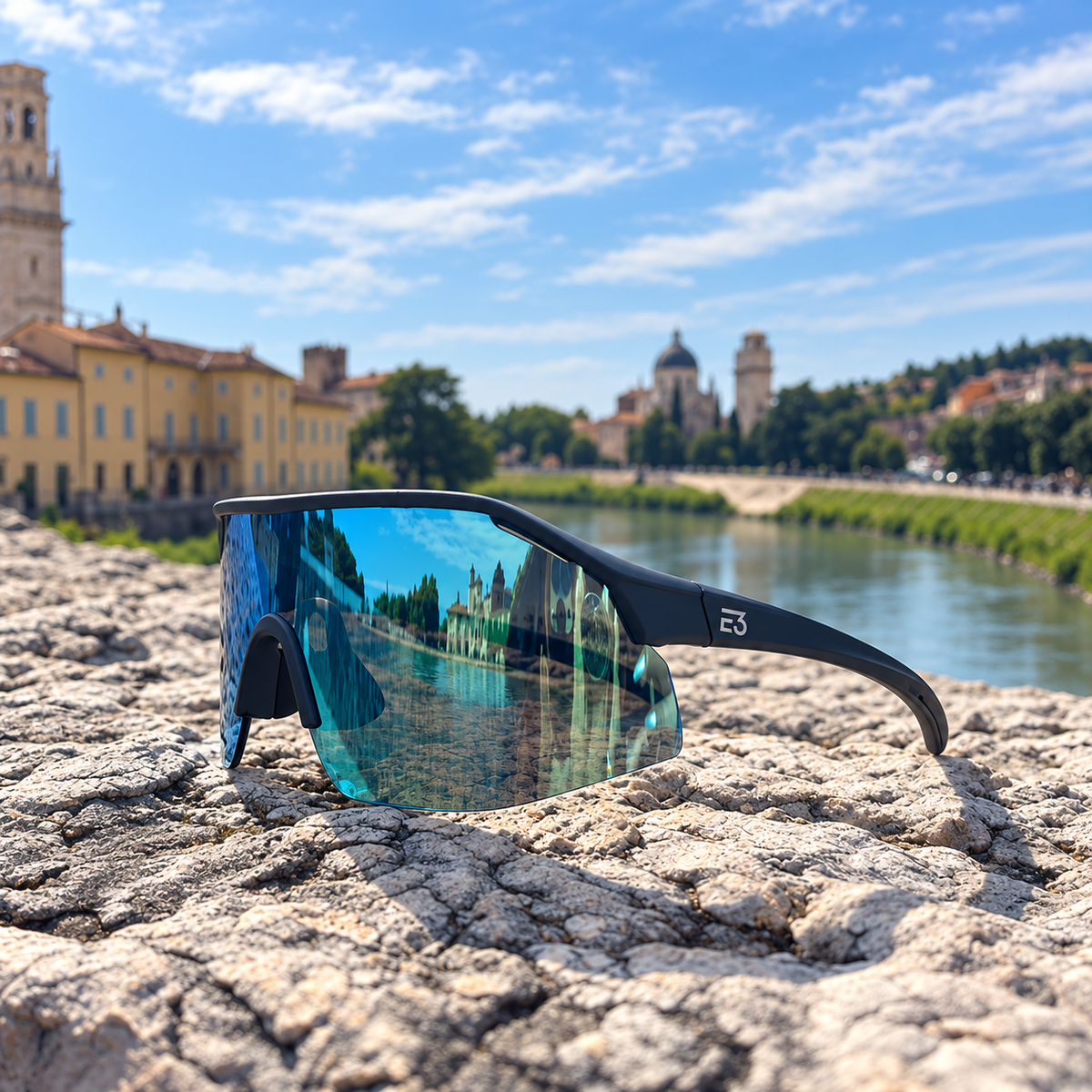 Black wrap-around sunglasses with a mirrored blue-green lens resting on a textured stone surface, with a scenic river and historic buildings in the blurred background.
