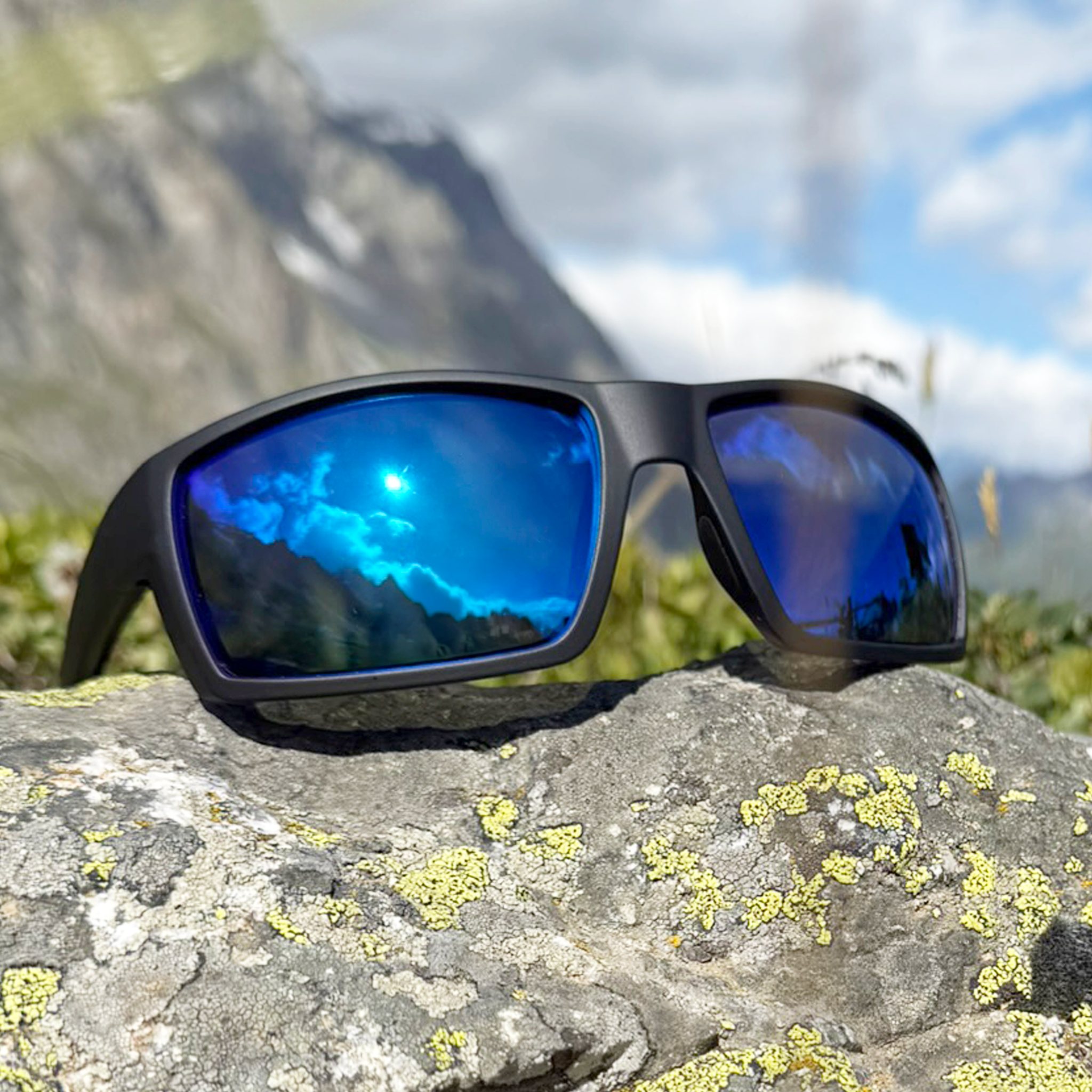 E3 Optics E2-A1 sport sunglasses with black frame and blue mirror polarized lenses, placed on a rock with mountains and sky reflected in the lenses