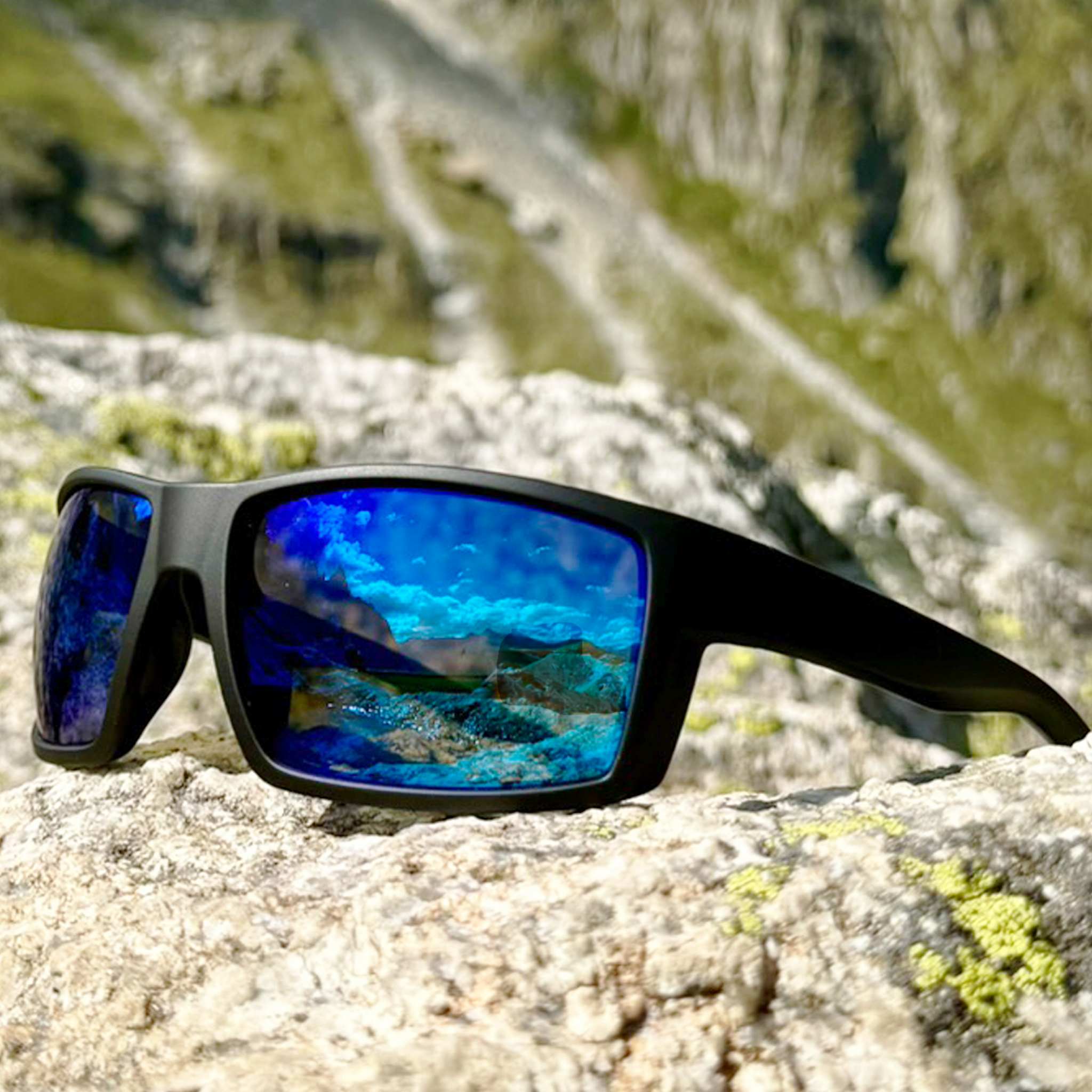 E3 Optics E2-A1 sport sunglasses with black frame and blue mirror polarized lenses, resting on a rock in a scenic mountain outdoor setting