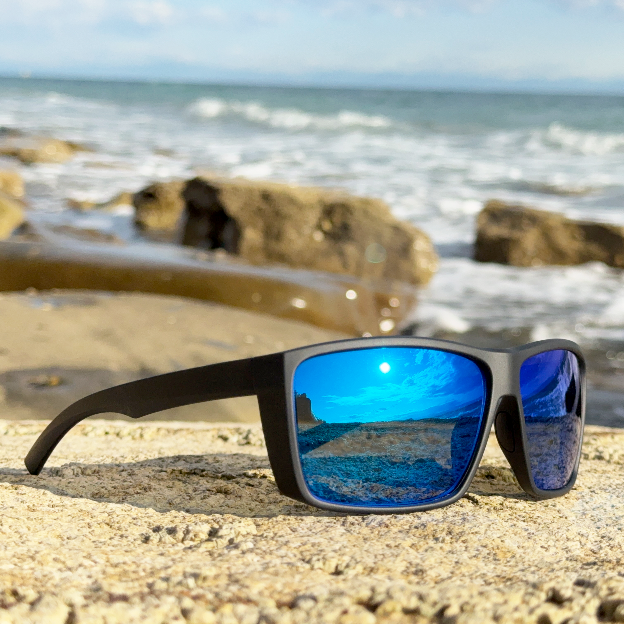 E3 Optics E1-B1 blue mirror polarized sunglasses with soft touch black frame – ideal for boating and fly fishing – right angle view on rocky beach with ocean in background
