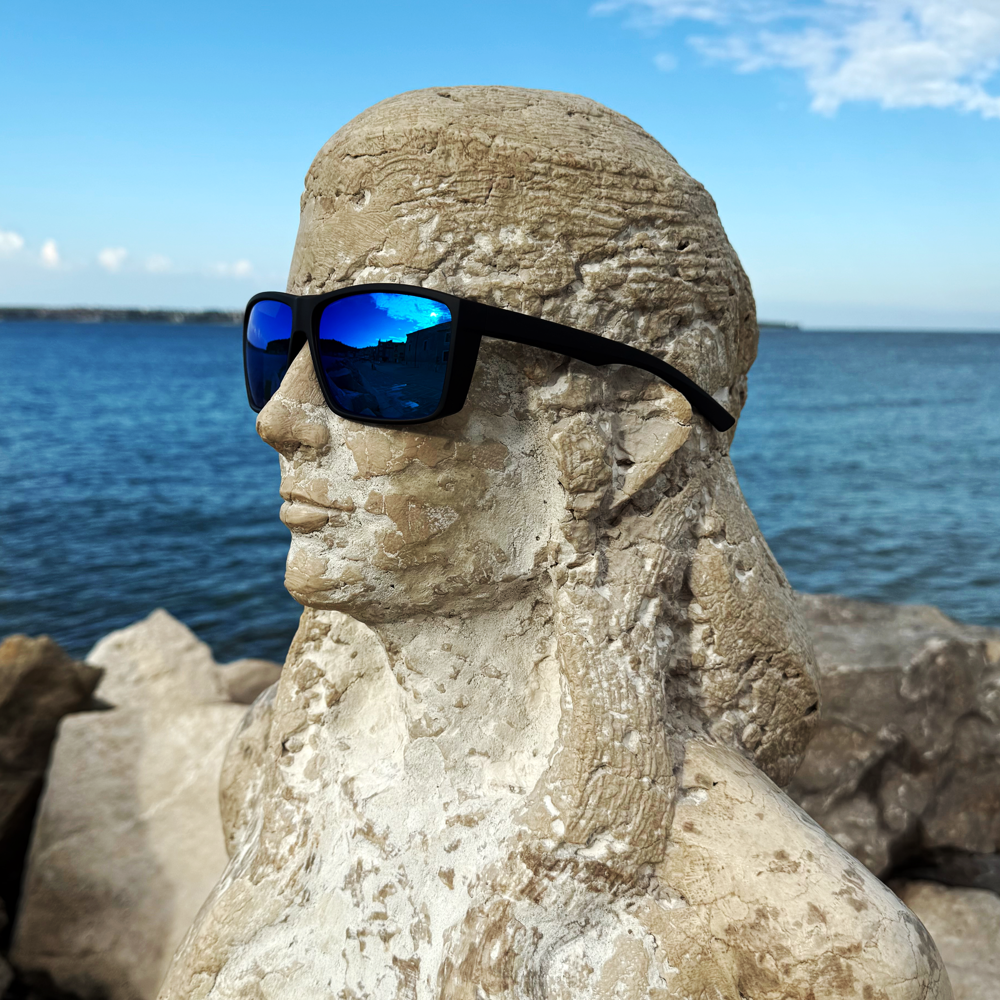 E3 Optics E1-B1 blue mirror polarized sunglasses with soft-touch black frame on a stone statue by the waterfront — ideal for boating and fly fishing — lifestyle side-profile view.