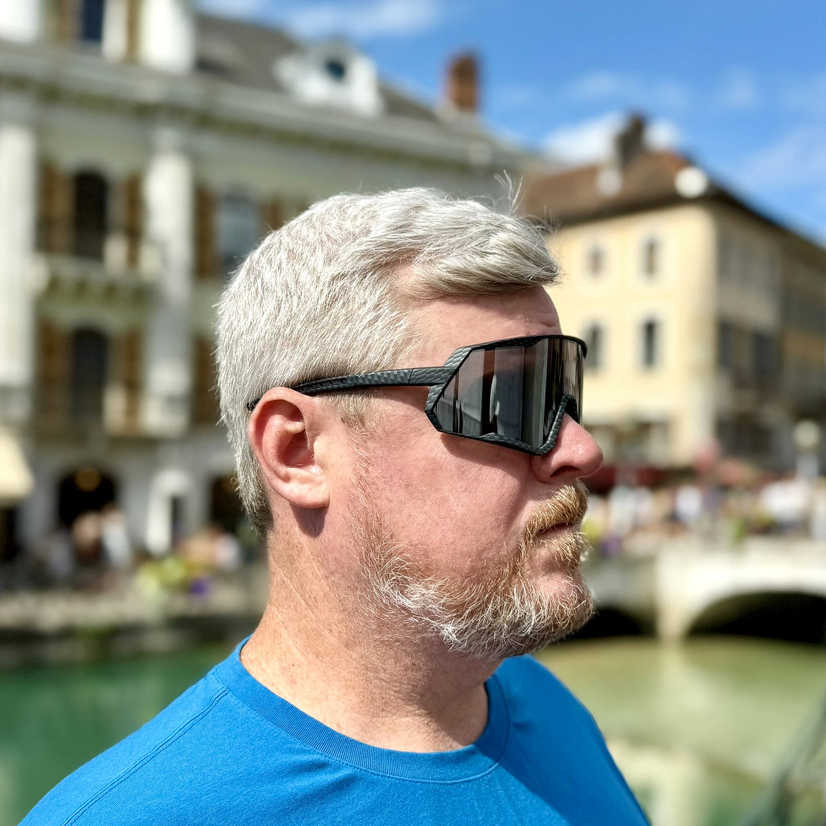 A man with gray hair and a beard wearing black wrap-around sunglasses and a bright blue shirt, photographed outdoors with a blurred canal and buildings in the background.