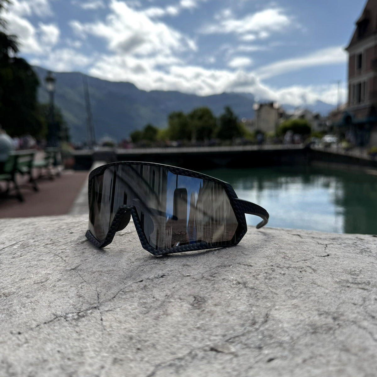Black wrap-around sunglasses with a dark mirrored lens resting on a stone ledge outdoors, with a blurred lakeside and mountain background.