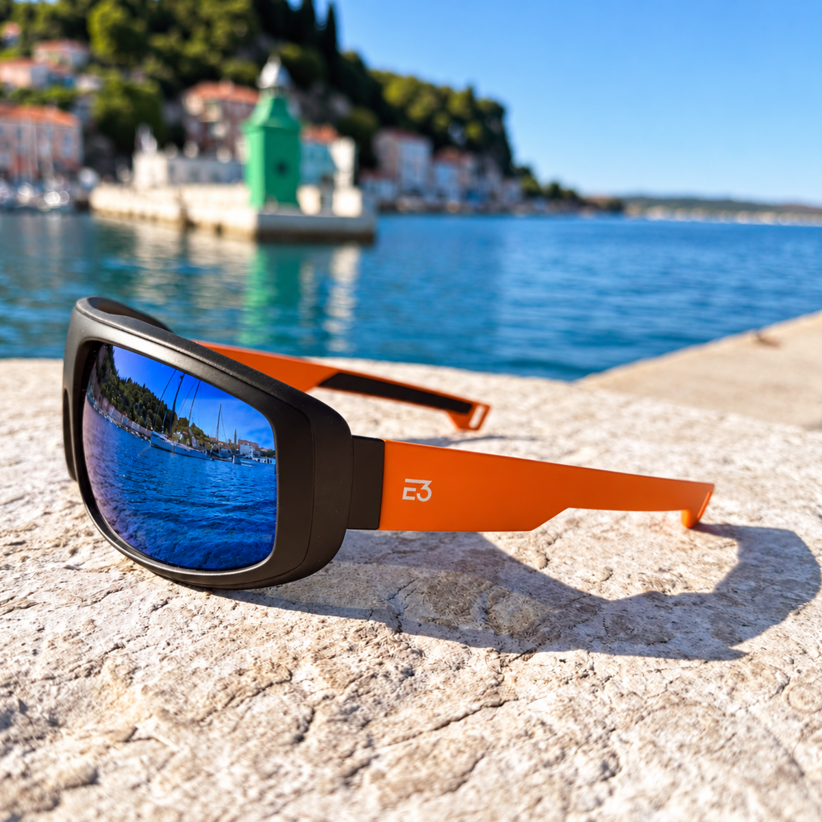 Orange and black sunglasses with blue mirrored lenses resting on a stone pier with a coastal harbor and buildings blurred in the background.