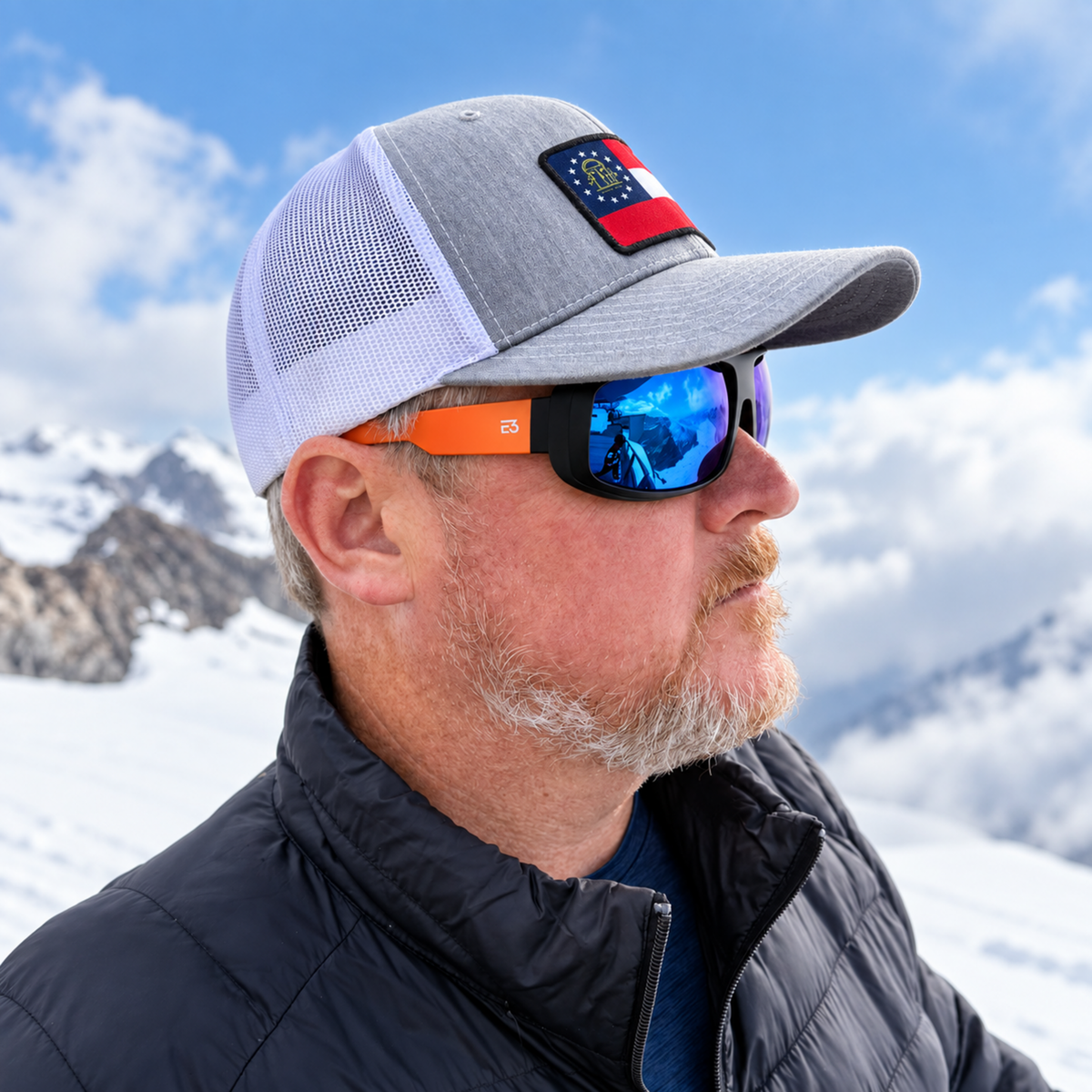A man in a snowy mountain setting wearing a gray and white trucker hat with a US flag patch, orange-framed sunglasses with mirrored blue lenses, and a black puffer jacket.