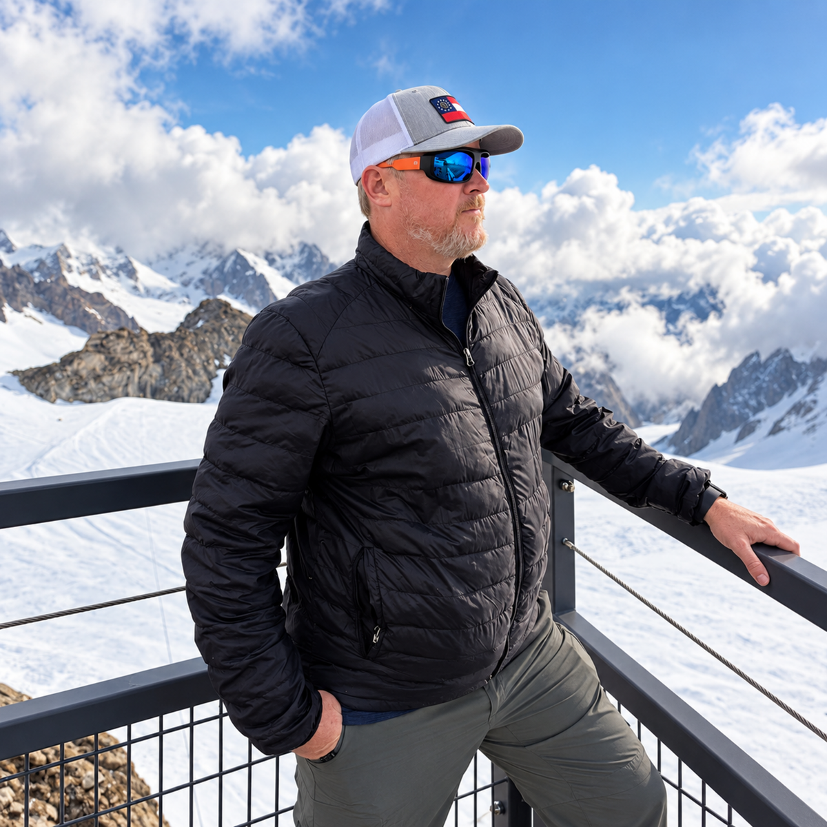 A man wearing a black puffer jacket, gray pants, a white and gray cap, and orange-tinted sunglasses stands on a balcony with a snowy mountain landscape in the background.