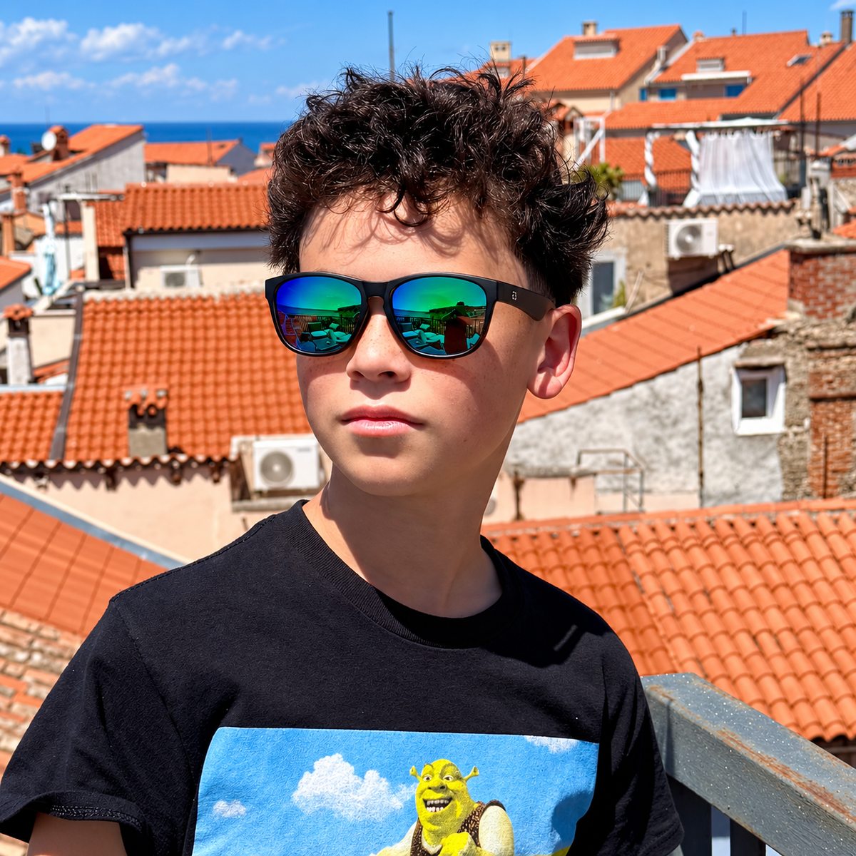 A young man with curly dark hair wearing black sunglasses with green-blue mirrored lenses and a black graphic t-shirt featuring Shrek, standing outdoors on a balcony with orange tiled rooftops and the sea in the background.