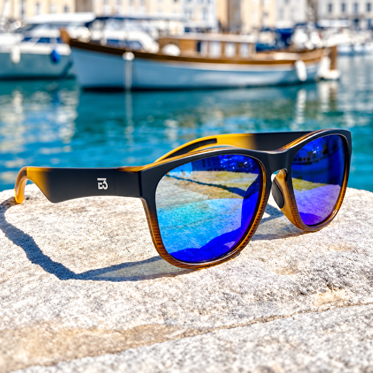 A pair of sunglasses with a black and amber tortoiseshell frame and vibrant blue mirrored lenses, resting on a stone surface with a blurred harbor and boats in the background.