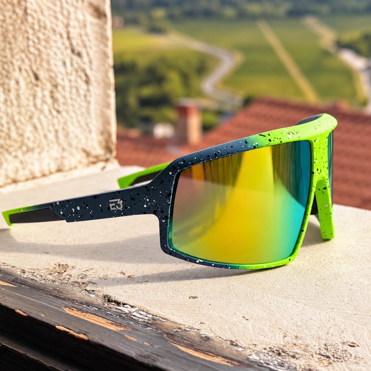 Sport sunglasses with a black and neon green speckled frame and mirrored yellow-green lenses, resting on a stone ledge with a blurred outdoor landscape background.