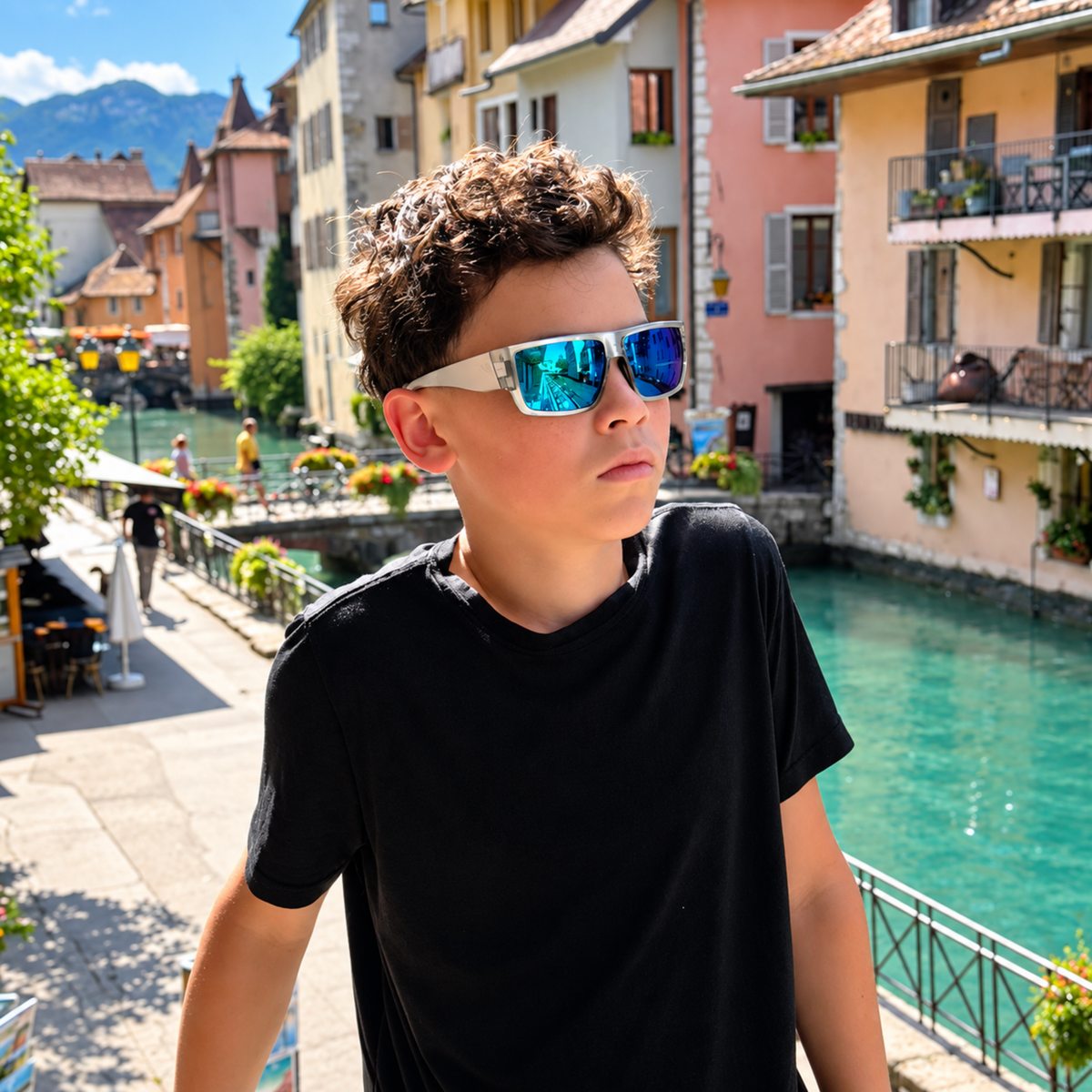 A boy wearing white-framed sunglasses with blue mirrored lenses, standing outdoors in front of a canal with colorful buildings and mountains in the background.