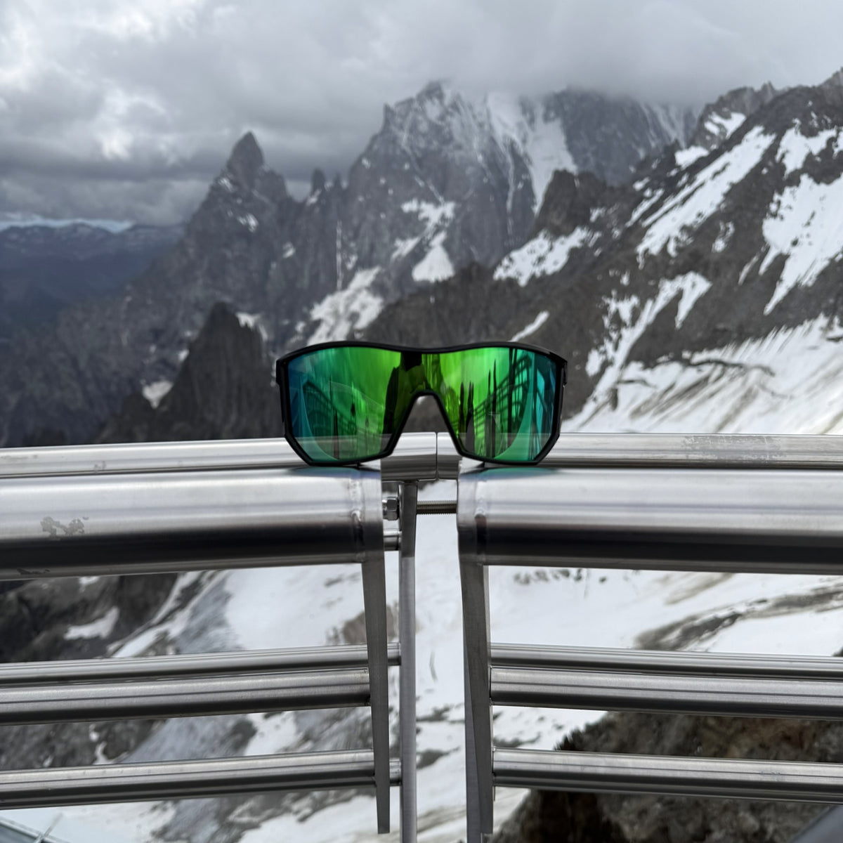 Black-frame E3 Optics Pulse sunglasses with a green mirrored lens resting on a railing above snowy mountains.