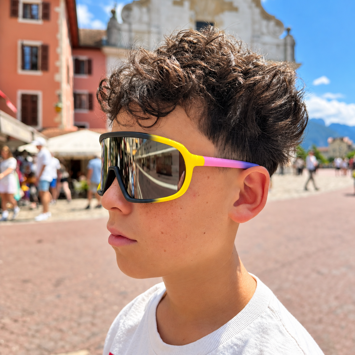 A boy in a white t-shirt wearing oversized shield-style sunglasses with a bright yellow frame, black upper rim, and a pink-to-blue gradient temple arm, photographed outdoors in a sunny town square.