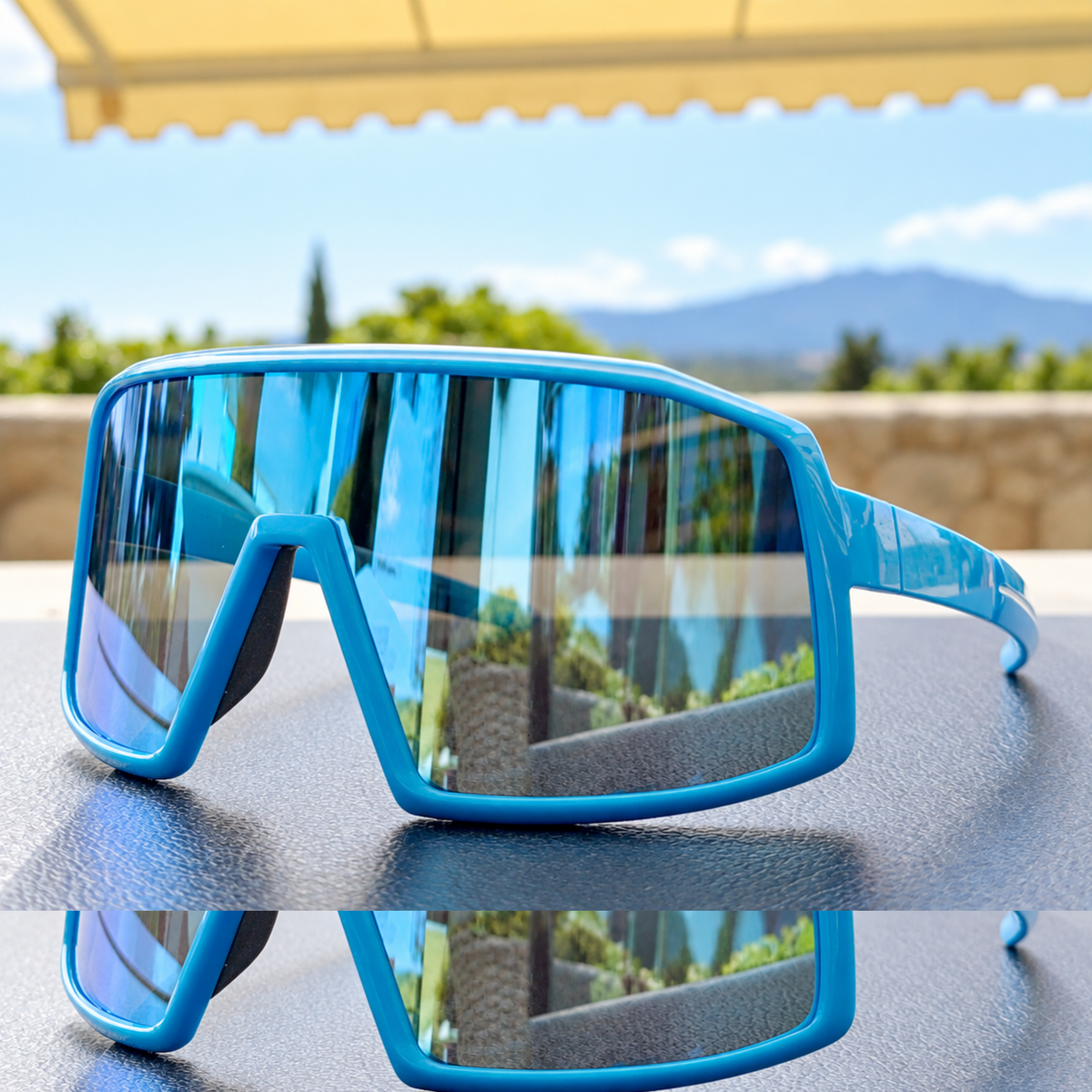 Blue wraparound shield-style sunglasses with mirrored lenses, resting on a dark textured surface with an outdoor patio and mountain background visible through the lenses.