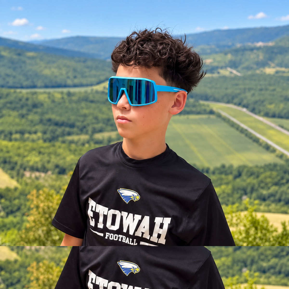 A boy wearing bright blue shield-style sunglasses and a black T-shirt with 'ETOWAH FOOTBALL' printed on the front, photographed outdoors against a blurred green mountain landscape.
