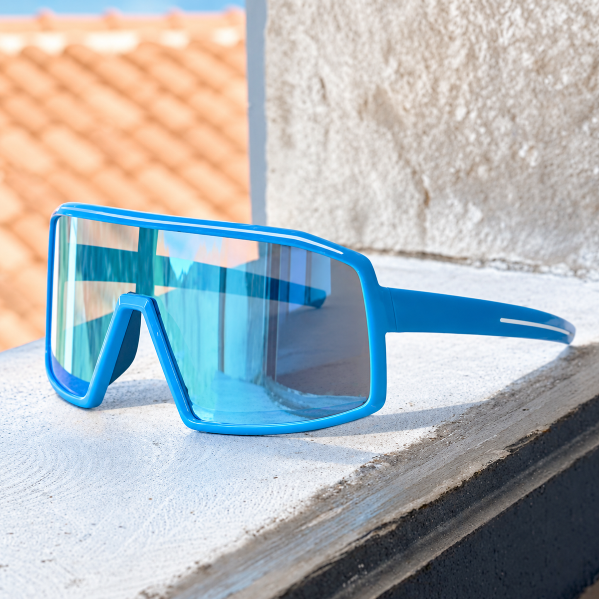 Blue shield-style sunglasses with a large wraparound lens and blue temple arm, resting on a white ledge outdoors with a textured wall and terracotta roof in the background.