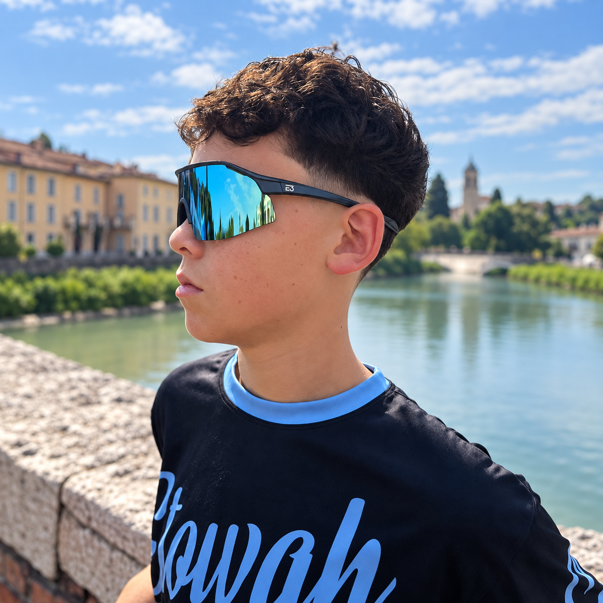 Side profile of a teen wearing blue mirrored E3 Optics Trace sunglasses beside the water, with waterfront buildings behind him.