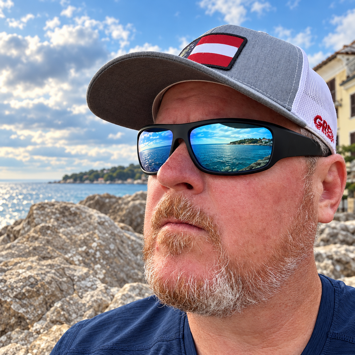 A man wearing a gray and white trucker hat with a red and white flag patch, black sunglasses with a blue ocean reflection, and a navy blue shirt, standing outdoors on a rocky beach with water and a building in the background.