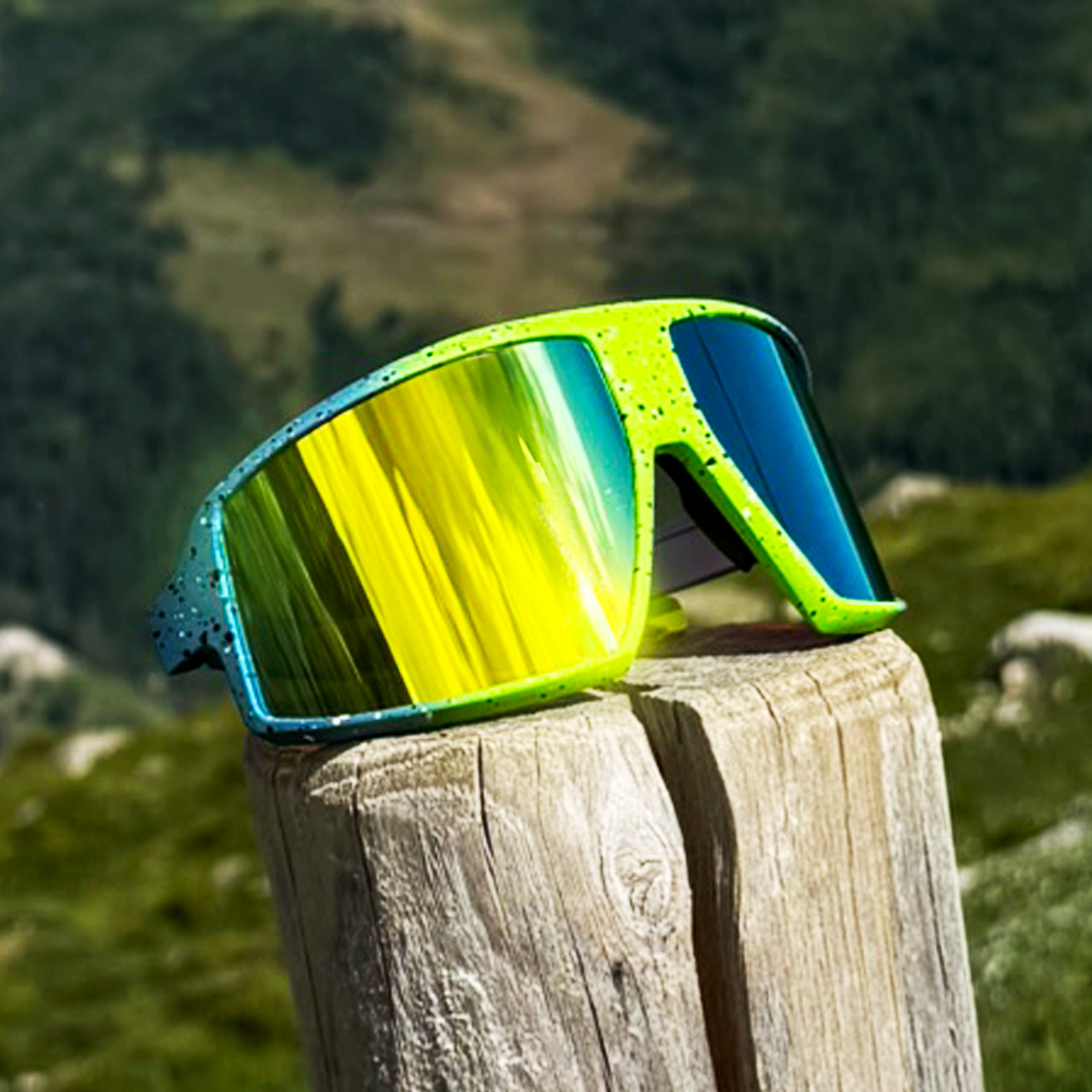 E3 Optics E9 precise lens sunglasses with green splatter frame resting on a wooden post in the mountains.